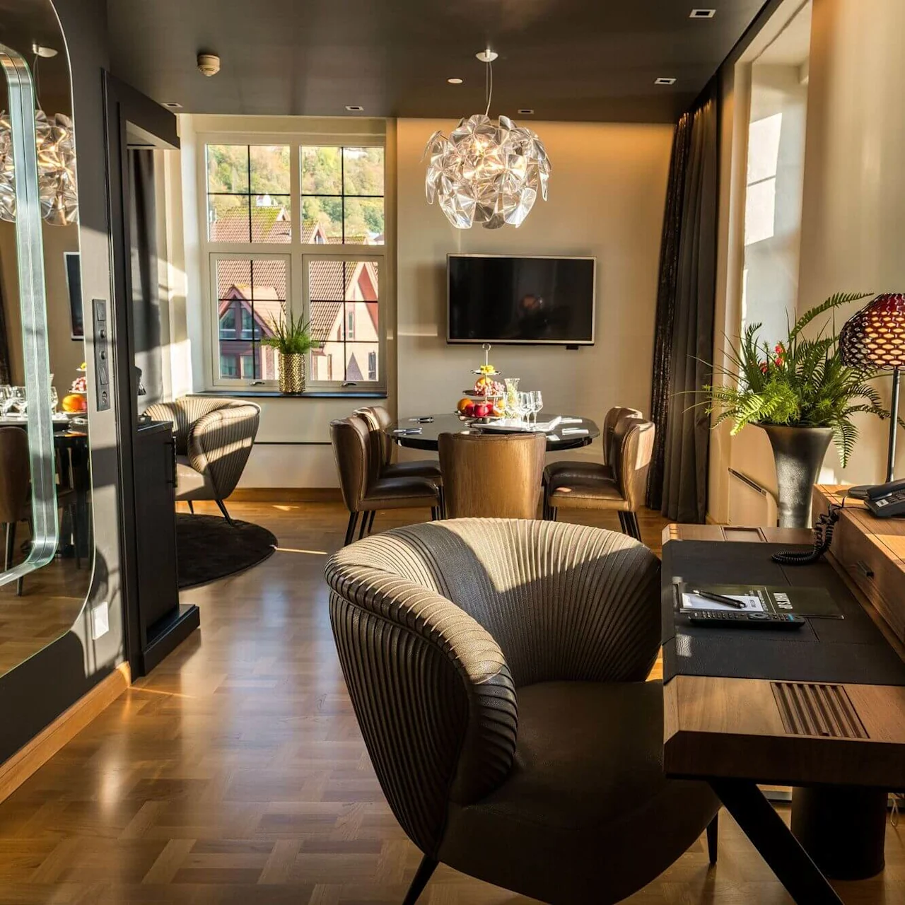 Interior of a stylish room with sunlight, featuring a dining table set, sofa, armchairs, and modern decor with a city view from a window.