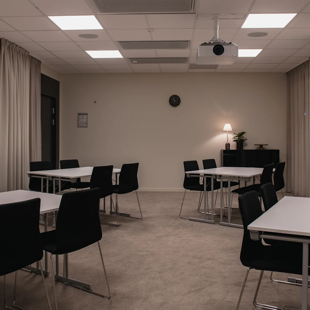 A classroom with white tables and black chairs arranged neatly. A projector hangs from the ceiling, and a clock decorates the wall. Curtains partially cover the windows.