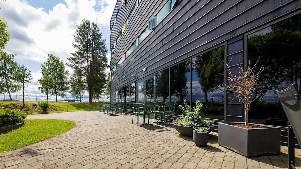 Rakennus, jossa on heijastavat lasi-ikkunat ja vihreitä tuoleja kivetetyllä patiolla, joka jatkuu nurmialueelle puiden keskellä pilvisellä taivaalla.