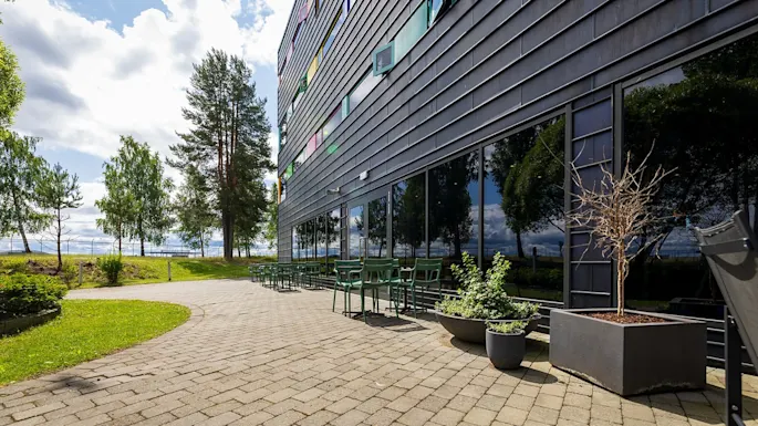 Building with reflective glass windows and green chairs on a paved patio extends into a grassy area with trees under a cloudy sky.