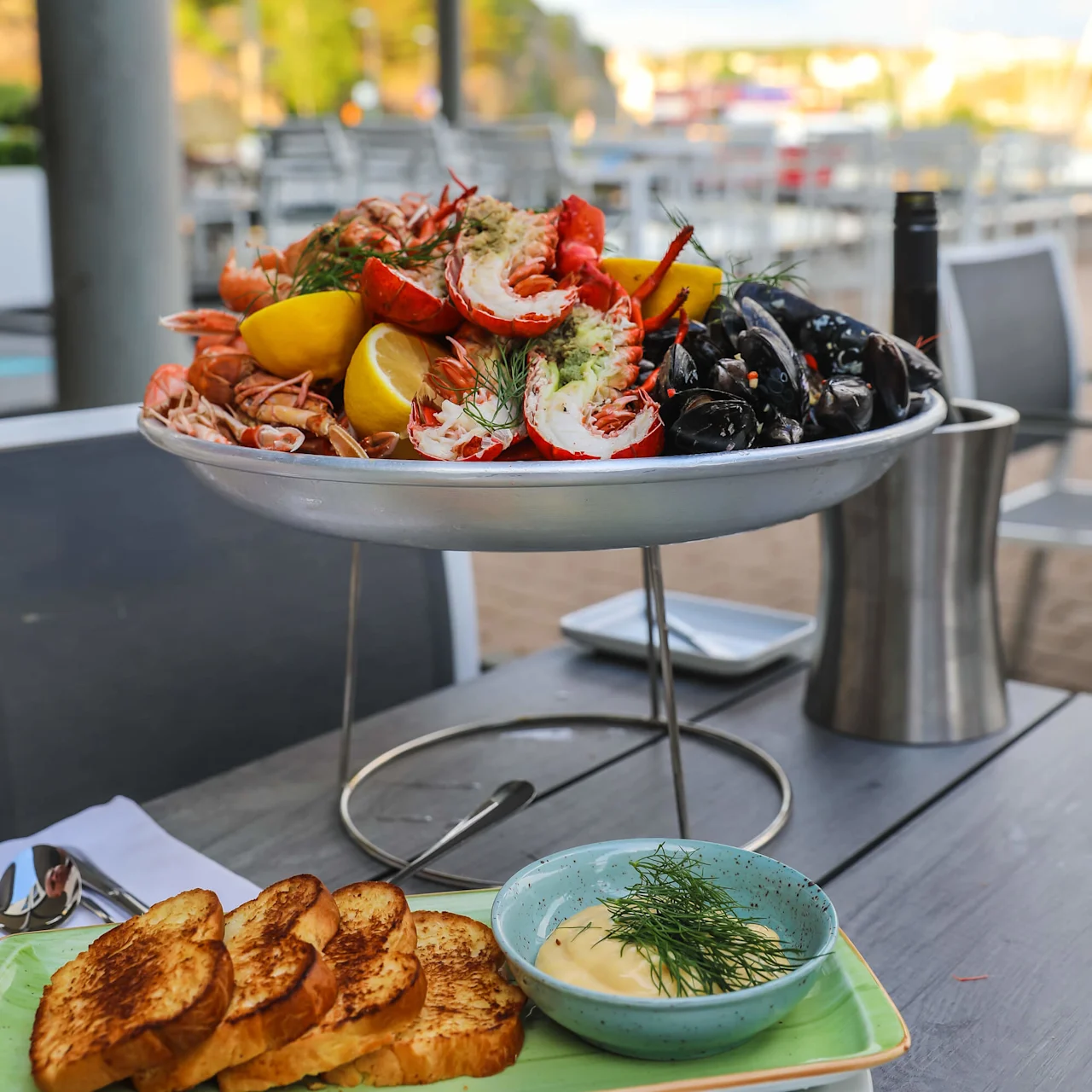 En tallerken med blandede skalldyr og sitronbåter er plassert på et utendørsbord med utsikt mot en uklar strandlinje. Ristet brød og en bolle med saus og urter ligger i nærheten.