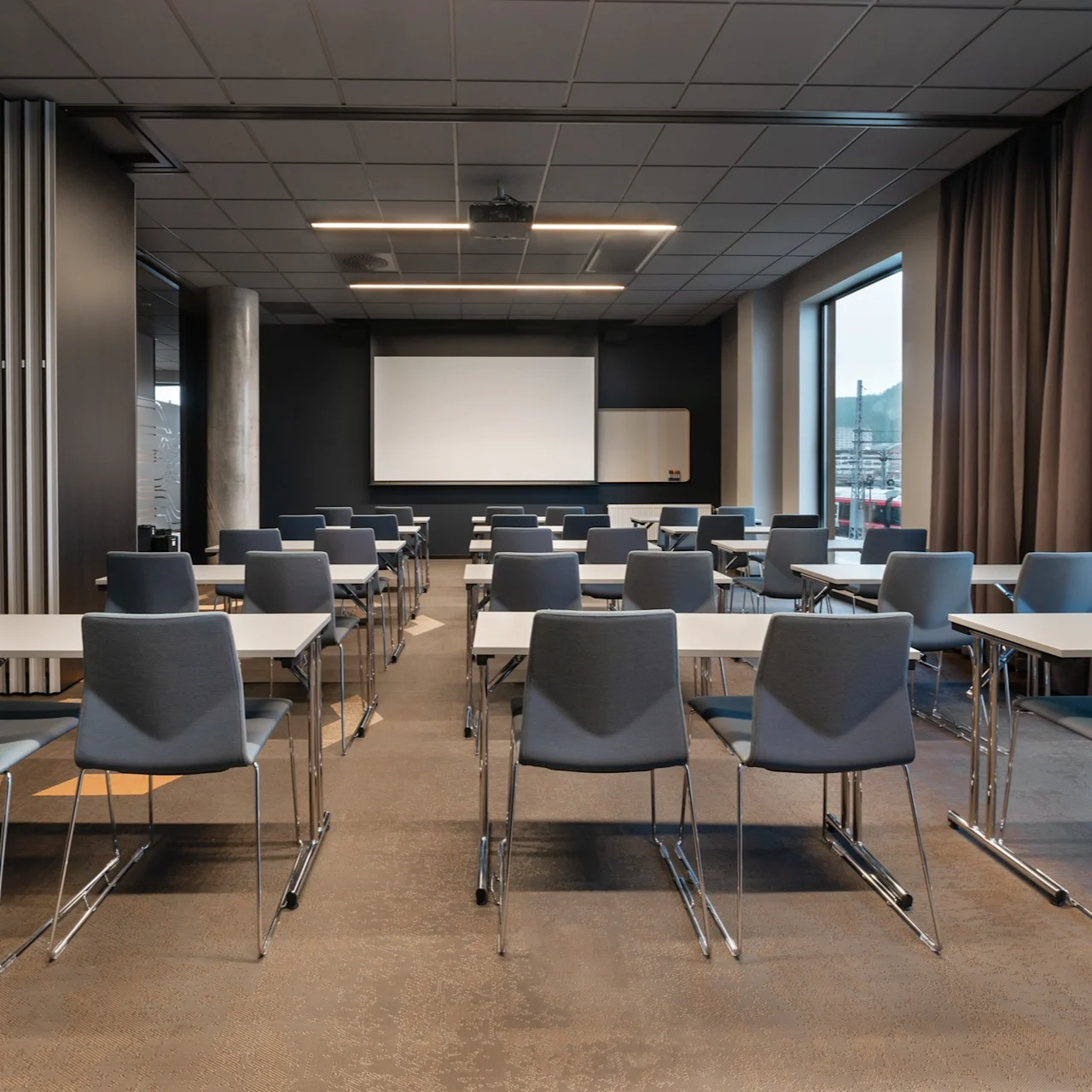 Rekker med tomme, grå stoler og hvite pulter vender mot en stor prosjektorskjerm i et moderne konferanserom; store vinduer og mørke gardiner er på den ene siden.