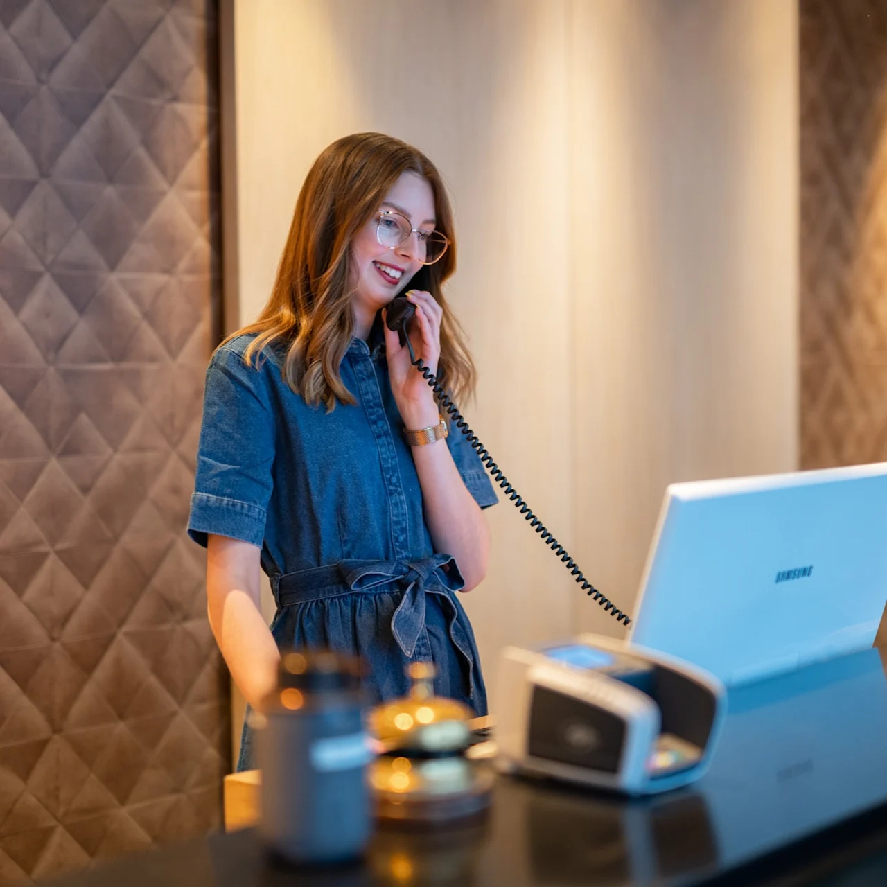 En person i en jeansklänning ler medan de pratar i en telefon med sladd vid en receptionsdisk. På disken finns en dator och kortläsare, med en quiltad vägg i bakgrunden.
