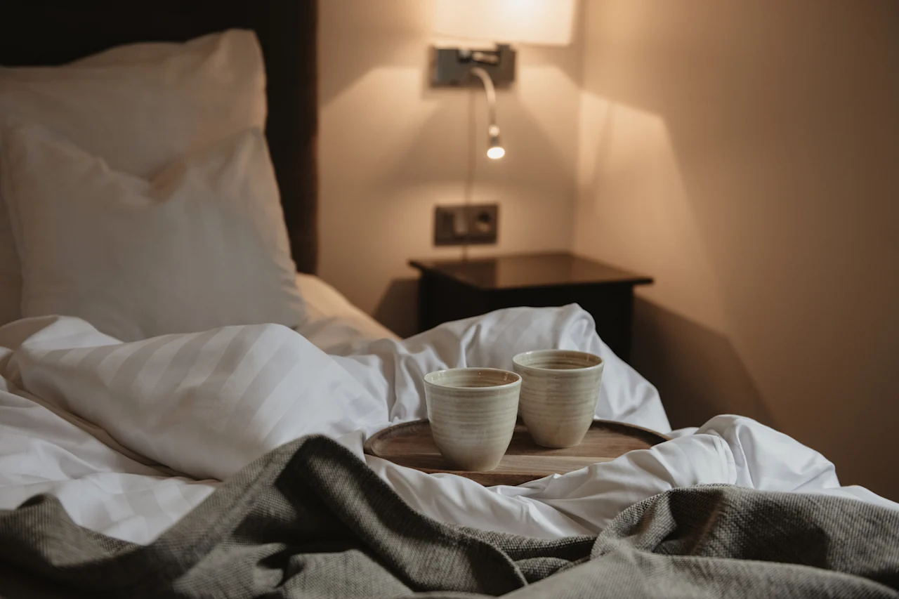 To keramiske kopper på en træbakke i en seng med hvide lagner og et gråt tæppe, hvilket antyder en hyggelig morgen på Home Hotel Norre Park i Halmstad.