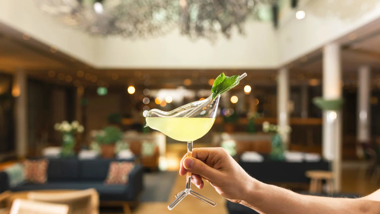 A hand presents a cocktail with a mint leaf garnish, blurred classy bar background.