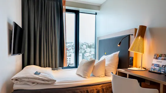 En nyredt seng med hvidt sengetøj og puder står ved siden af et skrivebord med en lampe og en brochure mærket "Tromsø." Et vindue viser udsigt over byens bygninger.