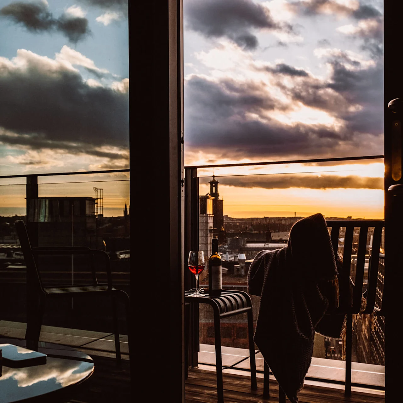 En stol, dekket med et teppe, har et vinglass og en flaske på en balkong under solnedgang. Bybildet strekker seg utover glassrekkverk under en dramatisk himmel.