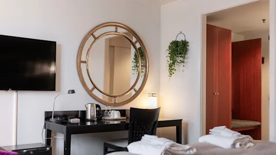 Viihtyisä hotellihuone, jossa on sänky puhtailla pyyhkeillä, työpöytä televisiolla ja koristeellinen pyöreä peili.