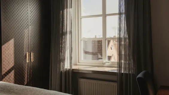 Ett mysigt hotellrum på Home Hotel Grand, Helsingborg, med solljus som strömmar in genom fönstret och lyser upp skira gardiner och en garderob.