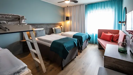 A hotel room with two single beds and a bunk bed occupies a light-blue painted room, featuring a ceiling fan, a window with curtains, a pink sofa, and a wooden floor.