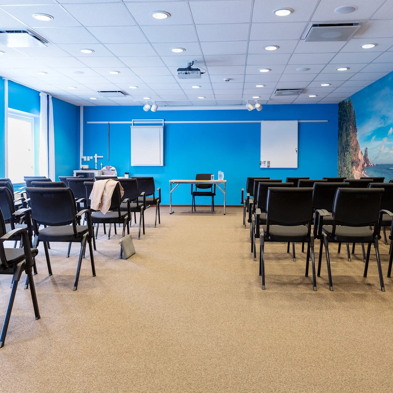 A spacious conference room with rows of empty black chairs facing a blue wall, featuring a flip chart and beach-themed mural, under bright ceiling lights. Windows line the left side.