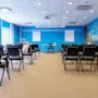 A spacious conference room with rows of empty black chairs facing a blue wall, featuring a flip chart and beach-themed mural, under bright ceiling lights. Windows line the left side.