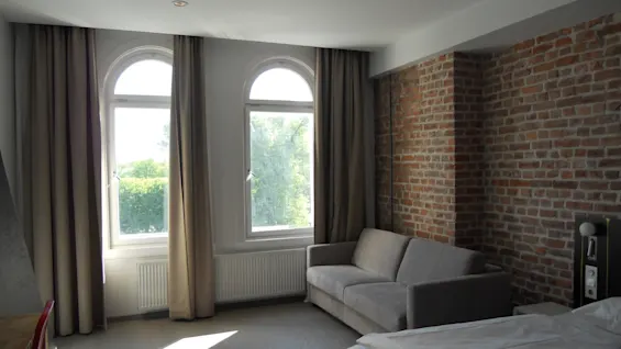A grey sofa sits against a brick wall, beside a bed in a room with wooden floors. Two large windows let in natural light with trees visible outside.