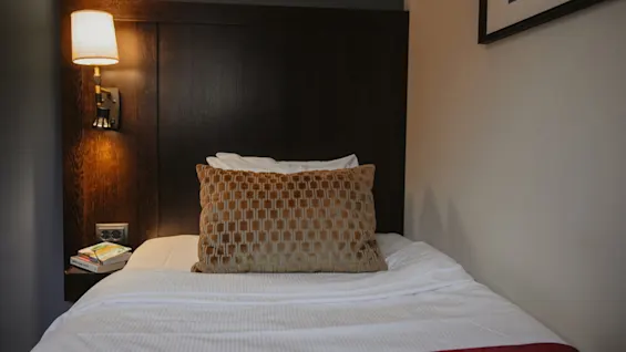 A cozy hotel room bed with a patterned pillow, white sheets, and a red throw, illuminated by a warm wall lamp. Home Hotel Kung Oscar.