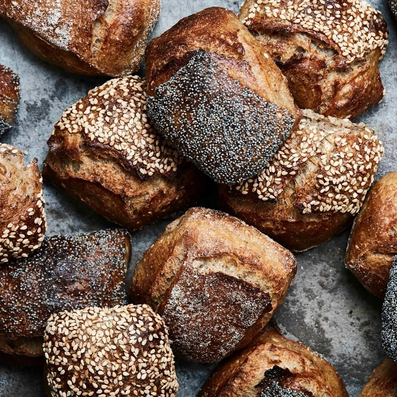 Bread rolls topped with poppy and sesame seeds lie scattered on a rustic, textured surface, showcasing their golden-brown crusts.