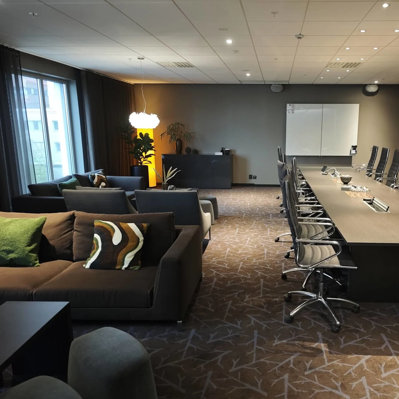Konferensrum med eleganta svarta stolar runt ett långt bord, intill en lounge med soffor och dekorativa kuddar. Fönster släpper in naturligt ljus över den moderna inredningen.