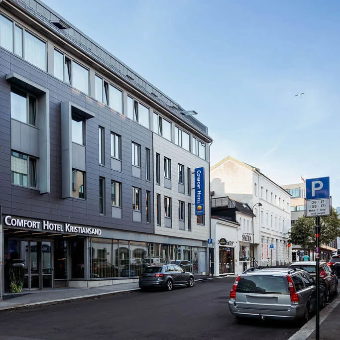 A modern hotel building labeled "Comfort Hotel Kristiansand" stands on a street with parked cars under a blue sky. A parking sign reads "P 08-17 (08 - 15)". 