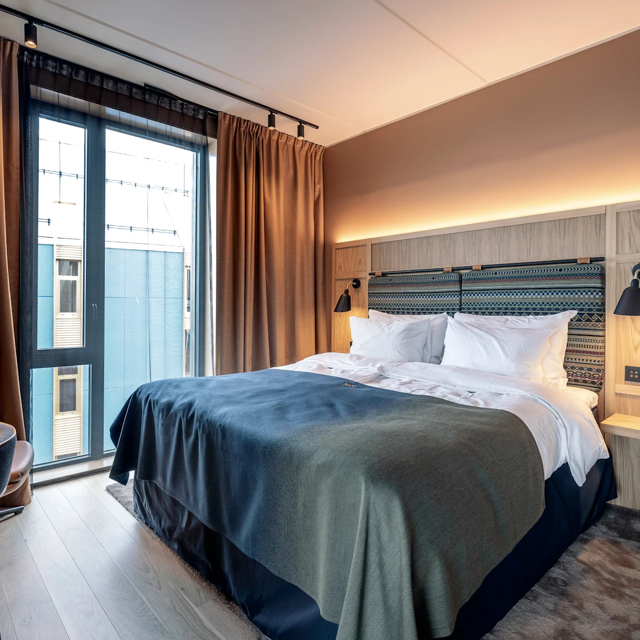 En seng med puder er centreret i et moderne hotelværelse, omgivet af et skrivebord, stol og et gulv-til-loft vindue med brune gardiner.