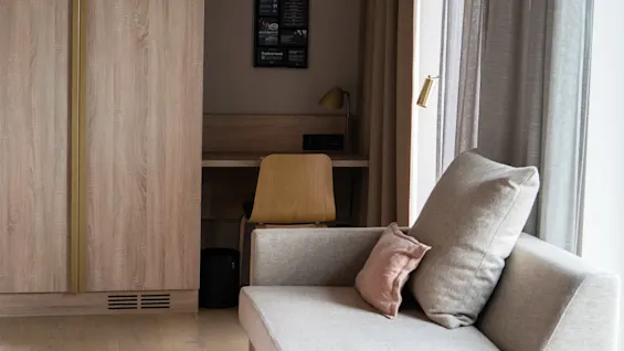 En beige sofa med puter står nær et vindu med grå gardiner. Bak den er et skrivebord i tre med en stol og lampe ved siden av et garderobeskap.