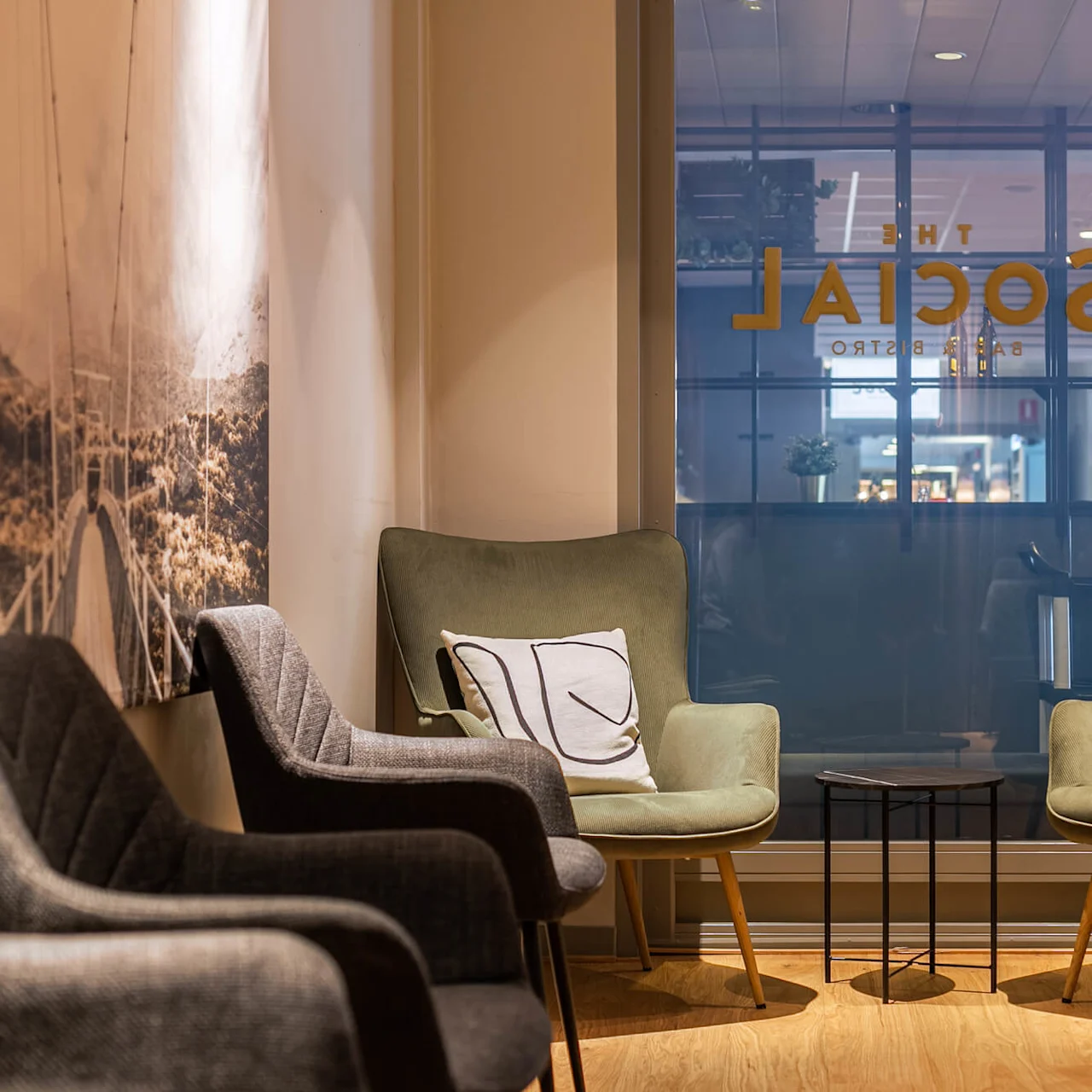 To grønne lænestole med dekorative puder flankerer et lille rundt bord. Et stort vindue med teksten "SOCIAL BAR & BISTRO" vender mod et lyst indendørs miljø. Flere stole står langs væggen.