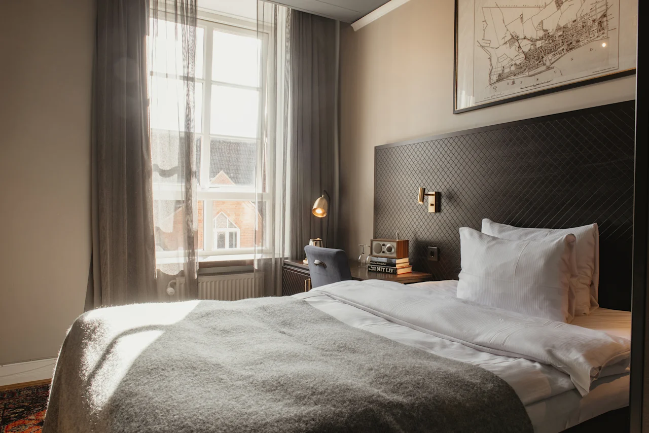 Ett solbelyst hotellrum på Home Hotel Grand, Helsingborg, med en bekväm säng och ett skrivbord vid fönstret. Text: MIT LIV.