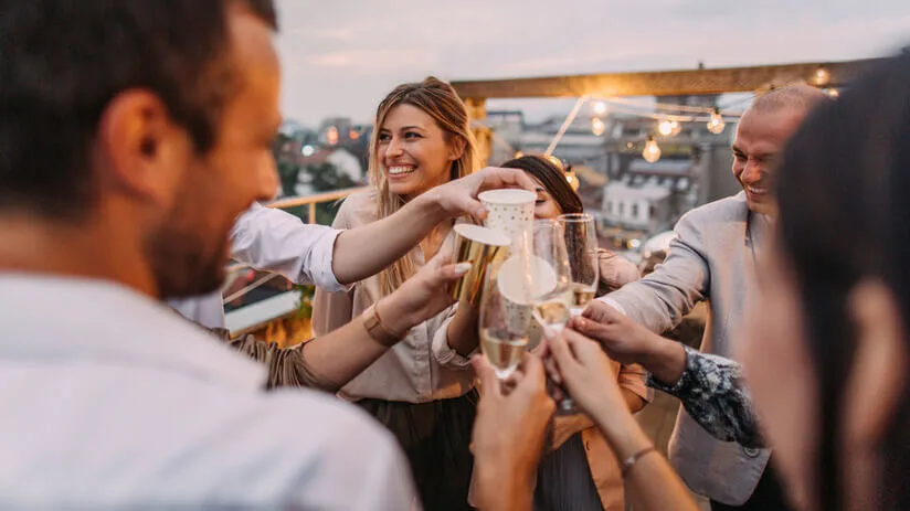 Grupp människor skålar med champagne på en takterrass i skymningen, stadssilhuett i bakgrunden, feststämning.