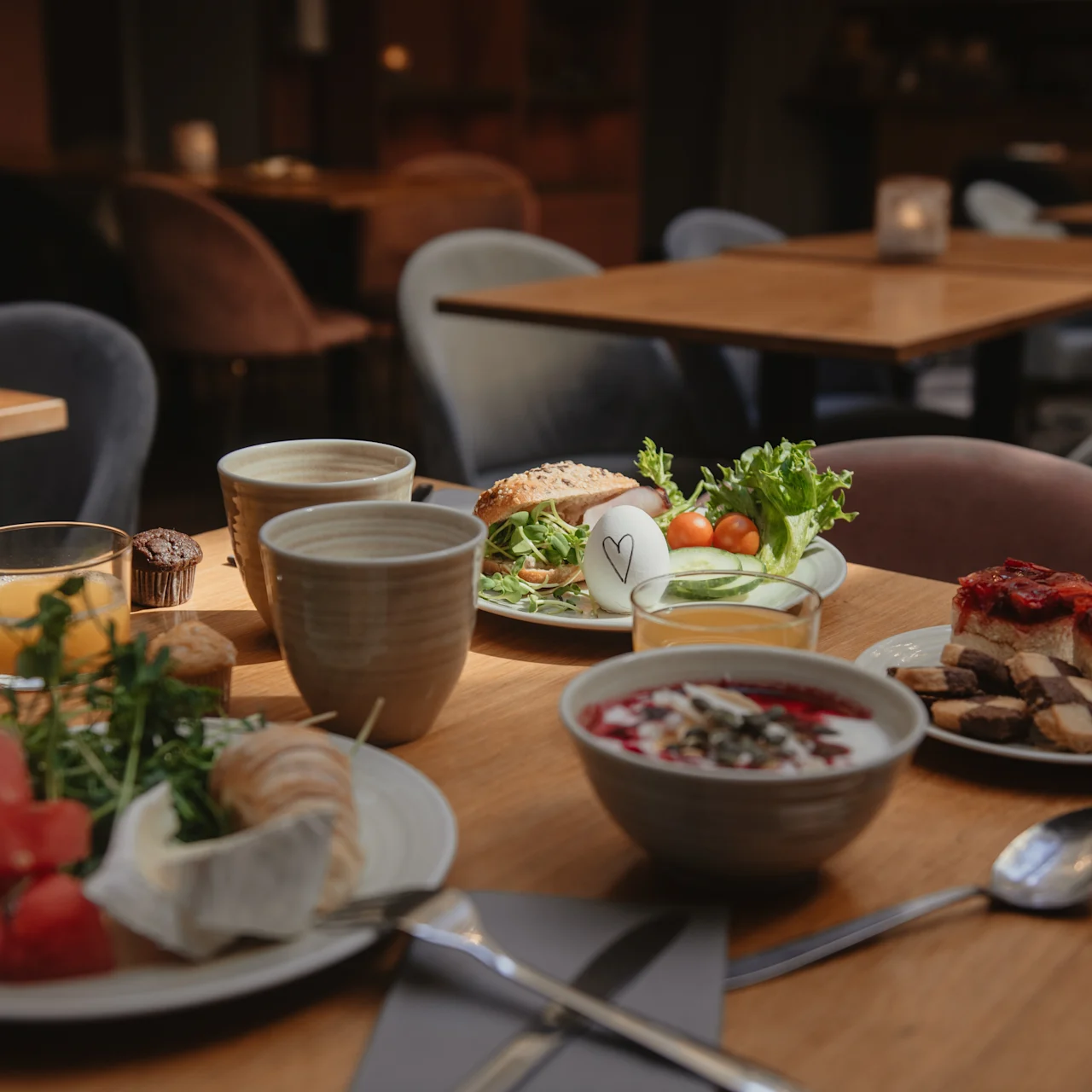 Et træbord er dækket med morgenmadsmad – frugt, yoghurt, sandwich og bagværk – omgivet af polstrede stole i et varmt oplyst spiseområde.