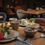 Ett träbord är dukat med frukostmat – frukt, yoghurt, smörgåsar och bakverk – omgiven av klädda stolar i ett varmt upplyst matområde.