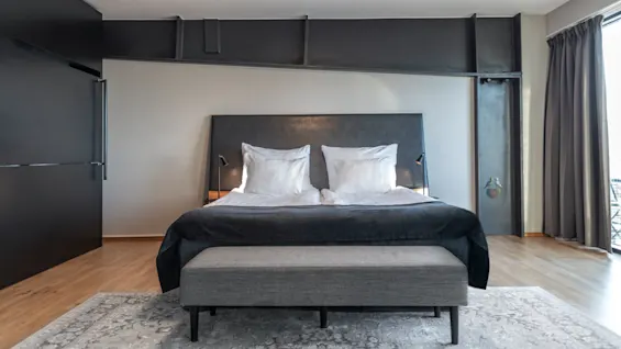 A neatly made bed with white pillows and dark covers stands in a modern bedroom with wooden floors, a gray rug, and large windows with dark curtains.