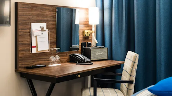 Ett hotellskrivbord med olika föremål, inklusive en telefon, hållare för anteckningsblock och en lampa, mot en bakgrund av blå draperi.