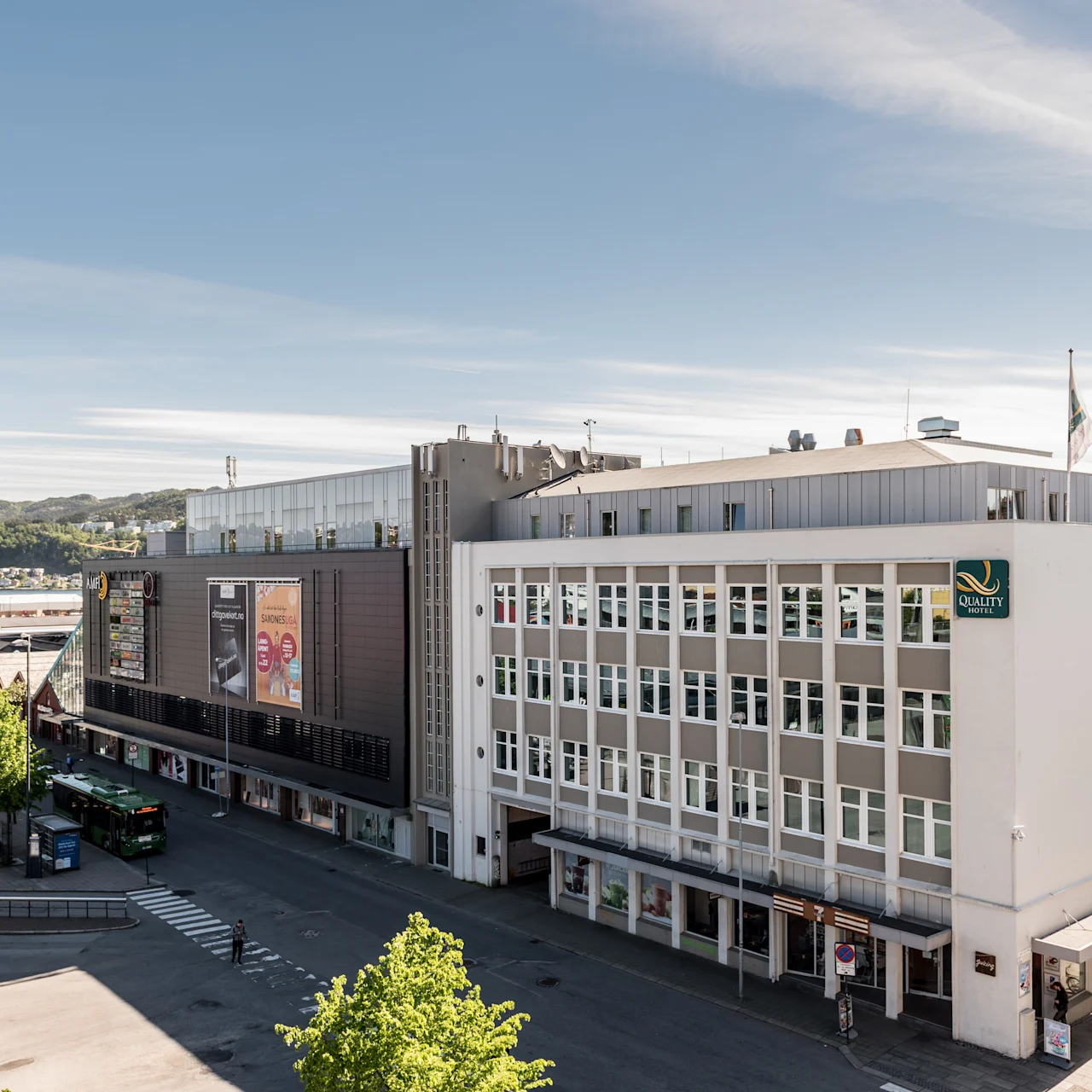 Usean kerroksen rakennuksessa on hotellin "QUALITY HOTEL" -kyltti ja lippuja katolla. Vihreä bussi on pysäköity edessä, aurinkoisena päivänä ja kirkas taivas.