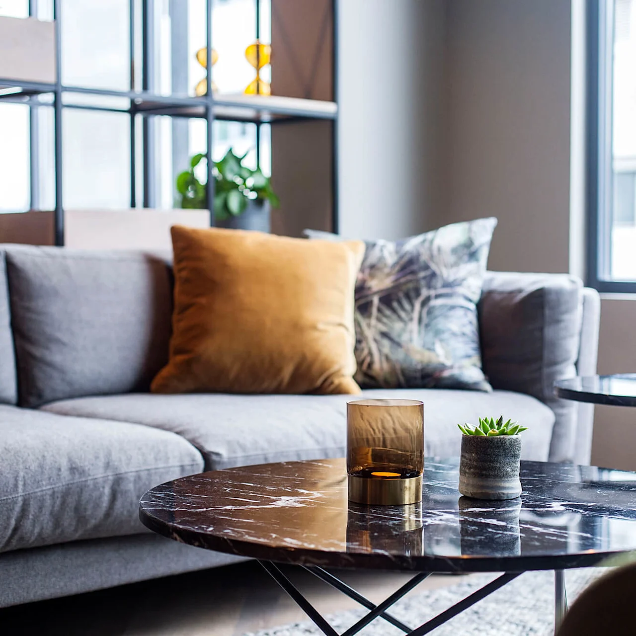 Grå sofa med sennepsgule og mønstrede puter, plassert i en moderne stue. Et marmorbord har en lysholder i glass og en liten plante, nær et vindu.