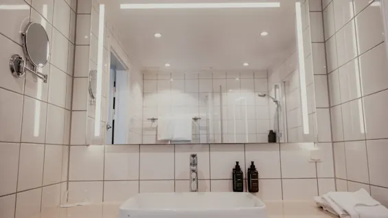 Ett rent och modernt hotellbadrum med en väl upplyst spegel ovanför ett handfat, med toalettartiklar och en fräsch handduk.