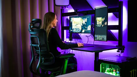 A person sits in a black ergonomic chair, gaming on dual monitors, illuminated by purple lights in a modern room with a mini-fridge containing drinks.