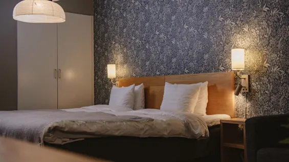 Bed with white linens in a cozy bedroom, flanked by two small wall lamps, against a patterned wallpaper backdrop. A wardrobe and chair are visible in the softly lit room.