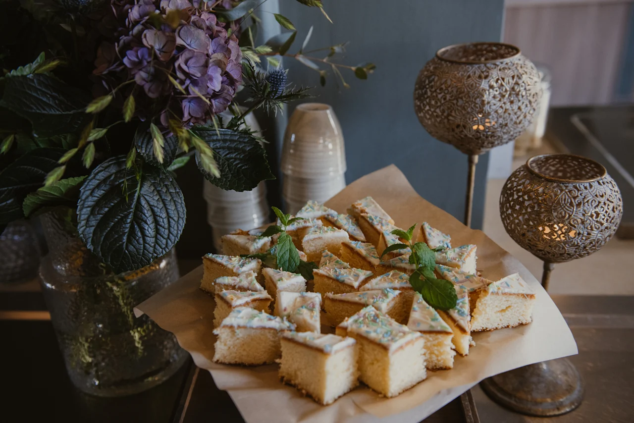 Ett fat med glaserade kakbitar med strössel och mynta, tillsammans med ett blomsterarrangemang och dekorativa lyktor, dukat för en sammankomst.