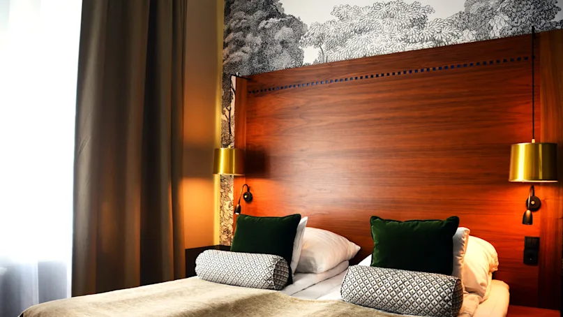 A cozy, neatly-made bed with pillows, flanked by brass wall lamps, against a wooden headboard and decorative wallpaper in a dimly lit room.