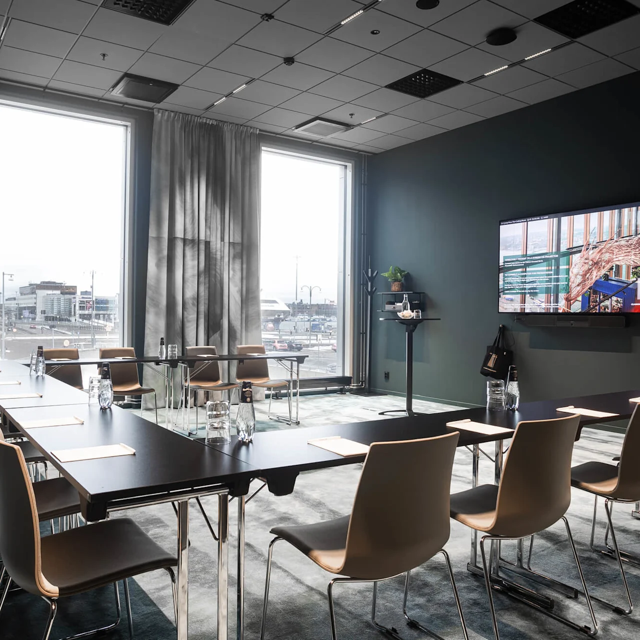 En konferensrum innehåller snyggt arrangerade stolar och bord, som vetter mot en väggmonterad skärm som visar en presentation. Stora fönster visar ett stadslandskap utanför.
