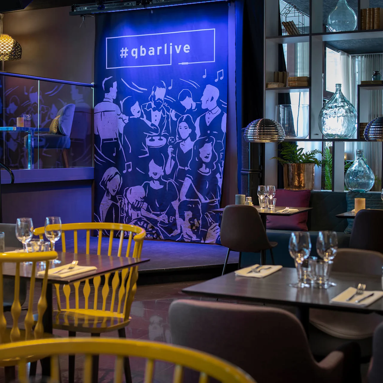 Middagsbord är dukade med glas och bestick i en stilfull restaurang. En scenbakgrund visar "#qbarlive" med konstnärliga figurer, omgiven av modern inredning och mjuk belysning.