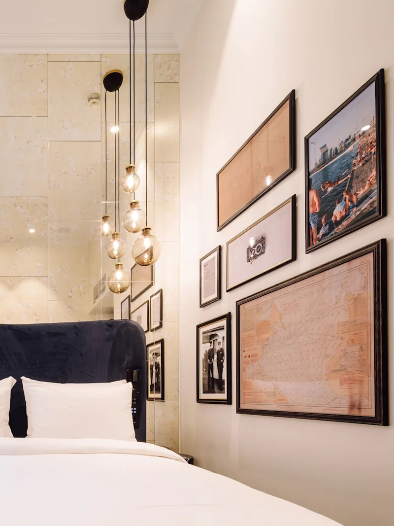 Sänky, jossa on valkoiset lakanat, on sijoitettu seinää vasten koristeltuna kehystetyillä kuvilla. Riippuvalaisimet roikkuvat yllä, suuret ikkunat ja verhot ovat vasemmalla.