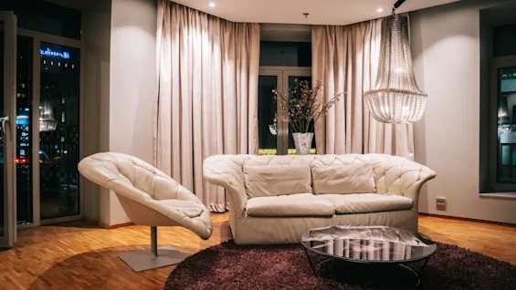 En mysig hotellounge på natten med gräddfärgad soffa, fåtölj, soffbord och ljuskrona. Stadsljus utanför. Text: edisson H.