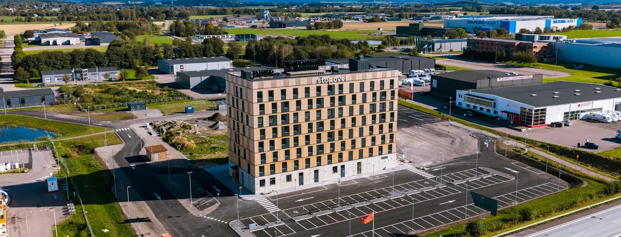 Ett högt, rektangulärt hotell med namnet "The Urban House" står på en stor parkeringsplats, omgiven av ett industriellt landskap med spridda lager och gröna fält under en delvis molnig himmel.