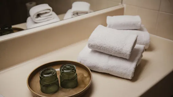 Two upside-down green glasses rest on a brown tray beside neatly stacked white towels on a beige bathroom countertop with a mirror reflecting them.