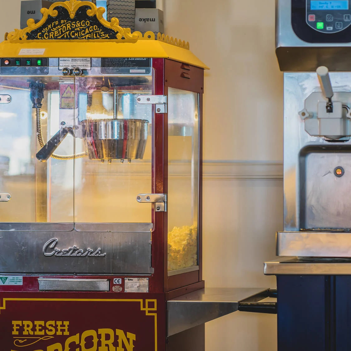 A red popcorn machine pops corn, situated in a kitchen with cups and supplies nearby. Text reads: "E. C. Cretors & Co., FRESH POPCORN."