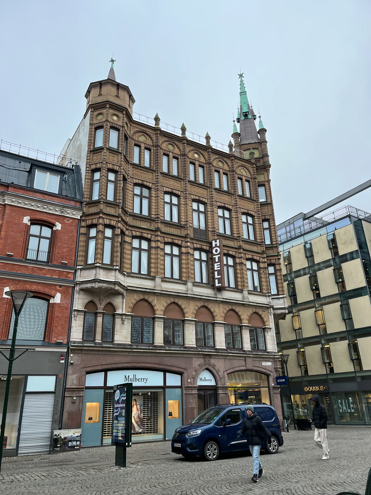 En fleretagers murstenshotelbygning med et grønt spir, med et "HOTELL"-skilt, står ved gaden, hvor folk går, og butikker, inklusive "Mulberry," kan ses nedenfor.