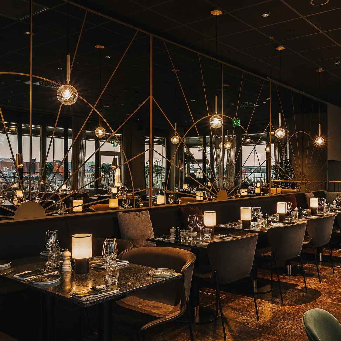 Spiseborde med glas og lamper, omgivet af polstrede stole, i en varmt oplyst restaurant med store vinduer, der giver udsigt til bybilledet. Elegante lysarmaturer hænger fra loftet.
