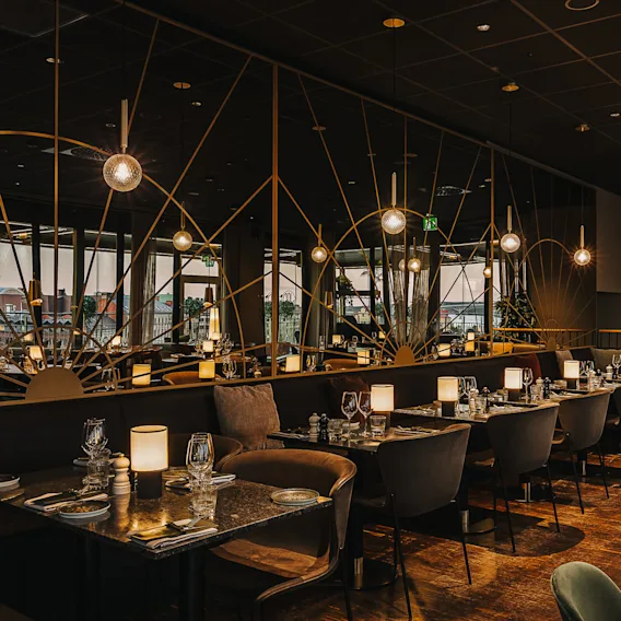 Dining tables set with glassware and lamps, surrounded by upholstered chairs, in a warmly lit restaurant with large windows overlooking a cityscape. Elegant lighting fixtures hang from the ceiling.