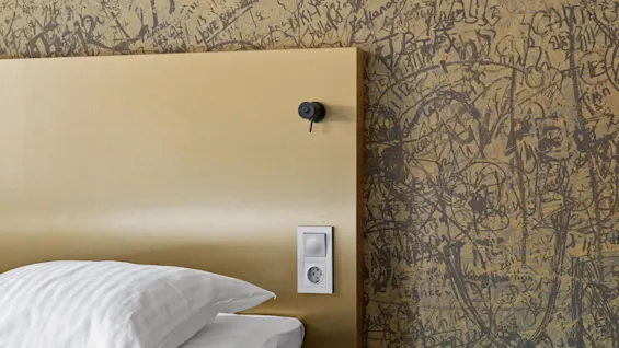 Seng med en hvid pude og beige sengegavl med et kamera og stikkontakt. Væggen er dækket af mørke graffiti-lignende kruseduller og tekst, inklusive "LOVE."