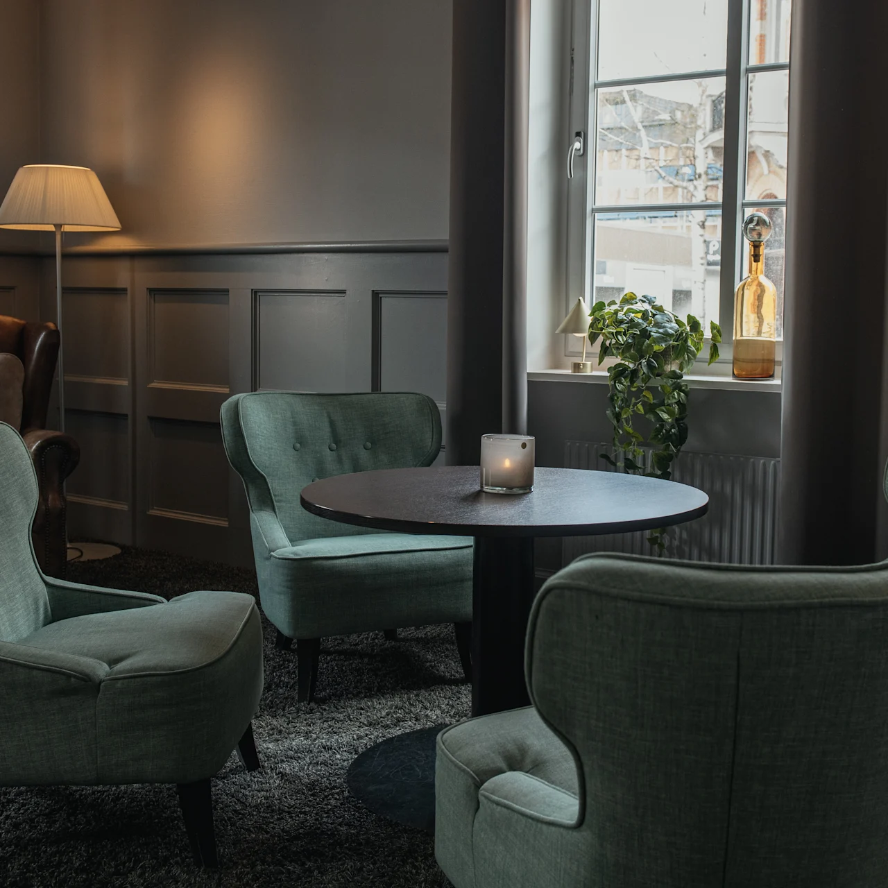 Four green armchairs encircle a round table with a candle, positioned in a serene room featuring a tall floor lamp and a window adorned with plants.