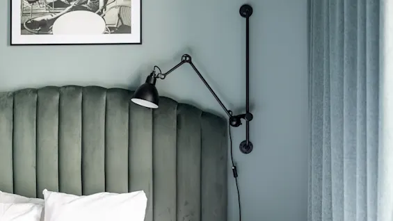 Et koselig soverom med en grønn fløyels hodegavl, hvite puter og en svart justerbar vegglampe. Et innrammet bilde av en utendørs kafé.